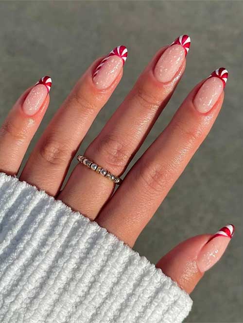 Almond-shaped red and white candy cane French tip nails on a nude base, adorned with white speckles resembling snow.