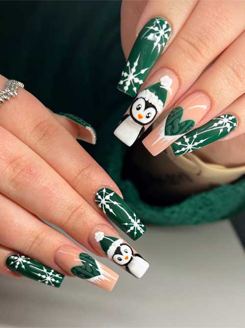 Dark green Christmas nails featuring snowflakes, with two nude accent nails adorned with a penguin and sweater heart designs.