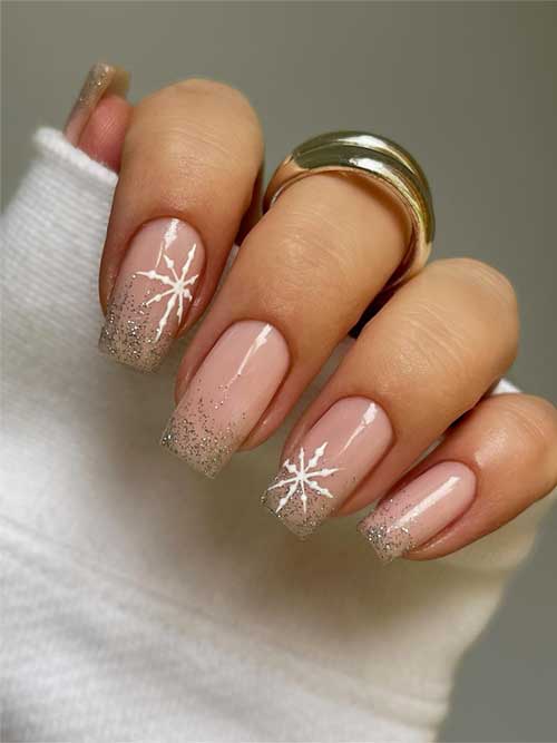 Square-shaped nude nails featuring gold glitter tips and two accent nails adorned with white snowflakes.