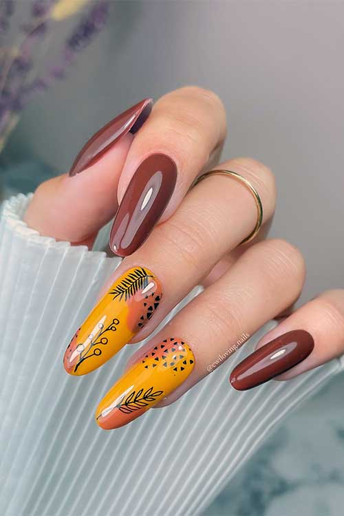 Glossy almond-shaped fall brown nails with two burnt yellow accents featuring black leaf and light brown abstract designs.