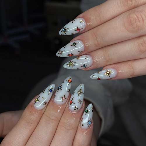 Long almond-shaped milky white nails adorned with gold stars and colorful rhinestones in red, blue, emerald, and pink.