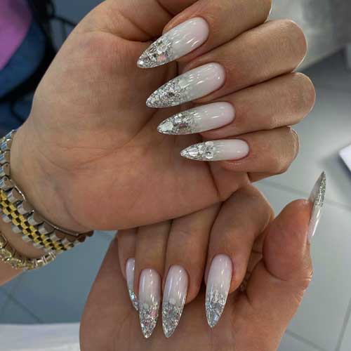 Long almond-shaped ombre New Year's nails transition from milky white to silver, adorned with large glitter flakes at the tips.