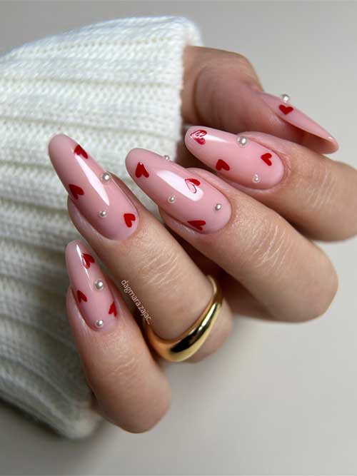 Glossy nude pink nails adorned with pearls and cute red hearts for a charming and stylish look.