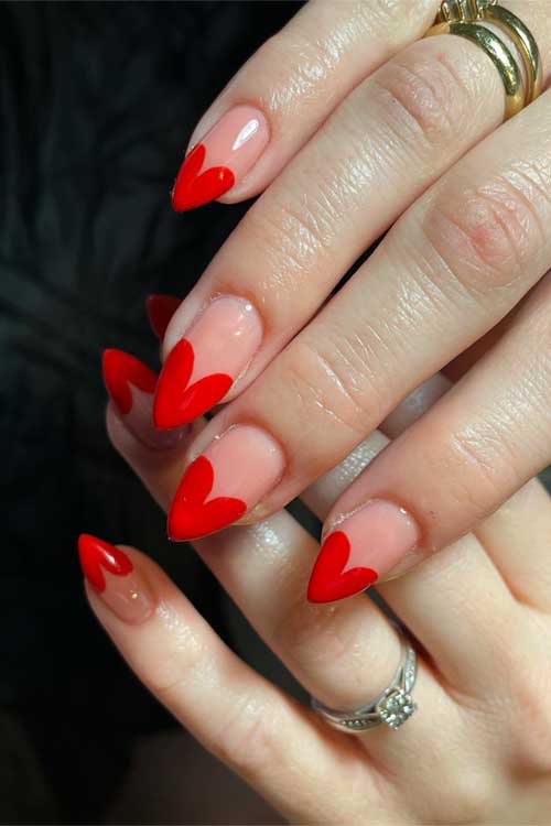 Nude pink almond-shaped nails featuring vibrant red heart tips for a playful and stylish look.