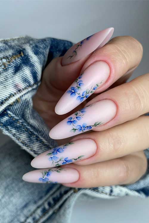 Long almond-shaped matte floral nails on a pale pink background, featuring delicate blue flowers and green leaves