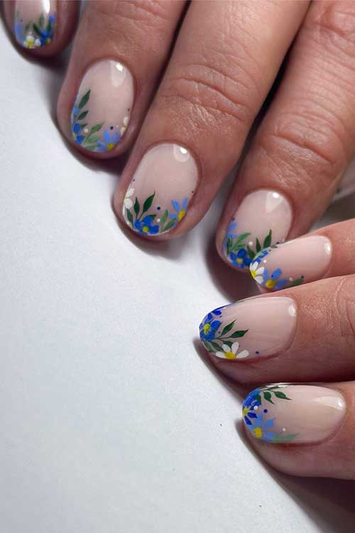 Soft nude nails with intricate blue and white floral patterns at the tips, accented by green leaves for a fresh spring look